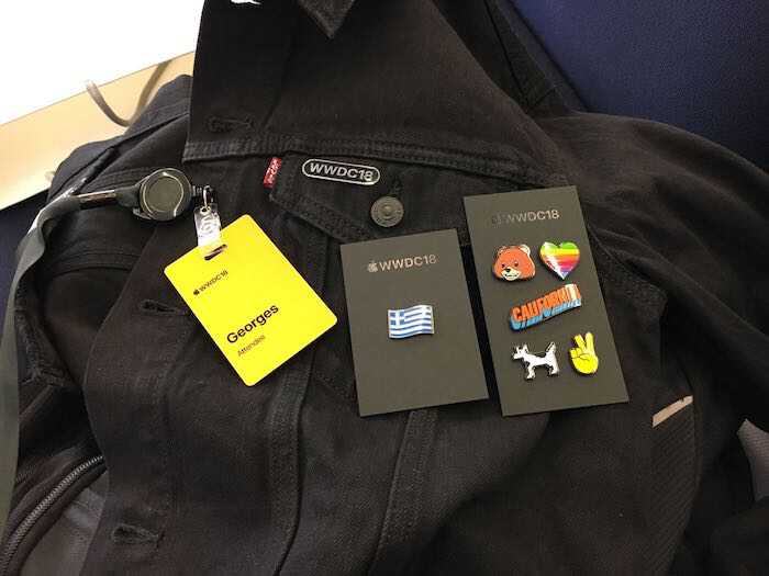 A photo of the WWDC18 black jacket with
									  layed out on top the yellow badge with
									  event's logo and the name George followed
									  by the word Attendee. There are two black
									  cards marked with AppleWWDC18 on top that
									  have colorful pins.  The first card has a
									  pin with the greek flag.  The second card
									  has six pins layed out in a rows of two
									  elements. The first row contains a bear
									  face and rainbow heart pin. The second row
									  contains the word California in a edged
									  design in orange and blue colors. The
									  third row contains the dogcow named Clarus
									  which is a bitmapped image designed by
									  Apple. The dogcow is black and white and
									  resembles a dog and a cow. The last pin is
									  a victory sign formed by the index and
									  middle fingers of a right hand.